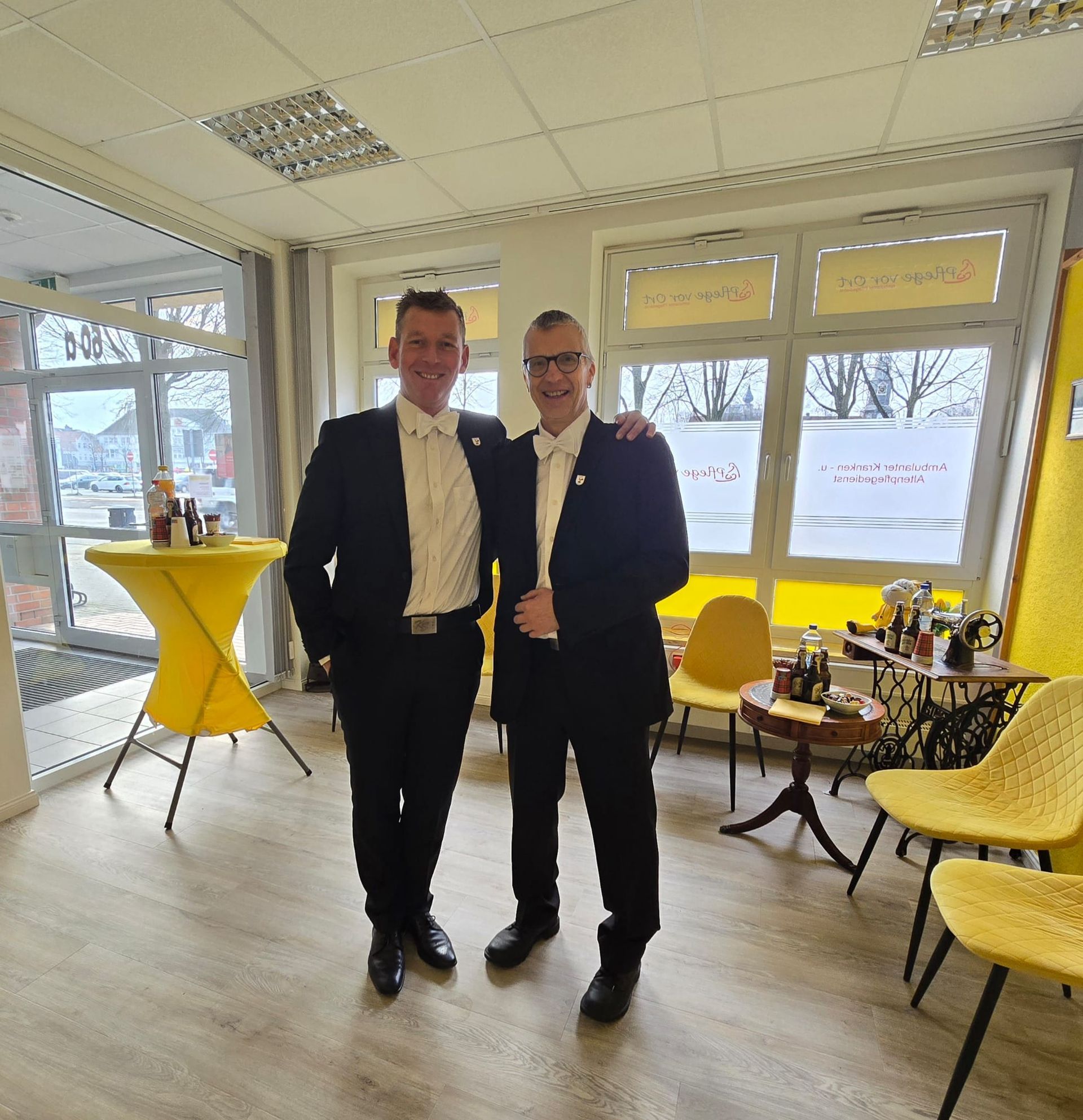 Zwei Männer in dunklen Anzügen stehen zusammen in einer hellen Lounge mit gelben Stühlen und hohen Fenstern.