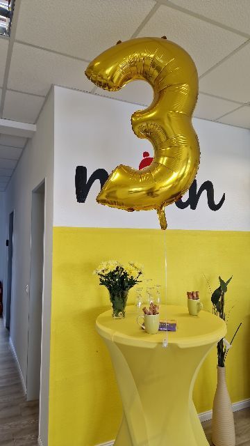 Ein goldener Ballon mit der Zahl 3 hängt über einem gelben Partytisch mit Blumen und Leckereien in einem hellen Raum.