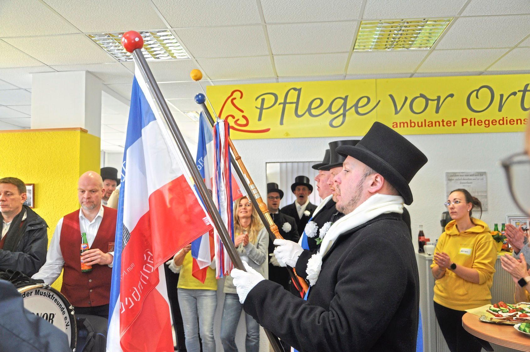 Menschen in Tracht halten Fahnen bei einem Indoor-Festival vor einem „Pflege vor Ort“-Banner.