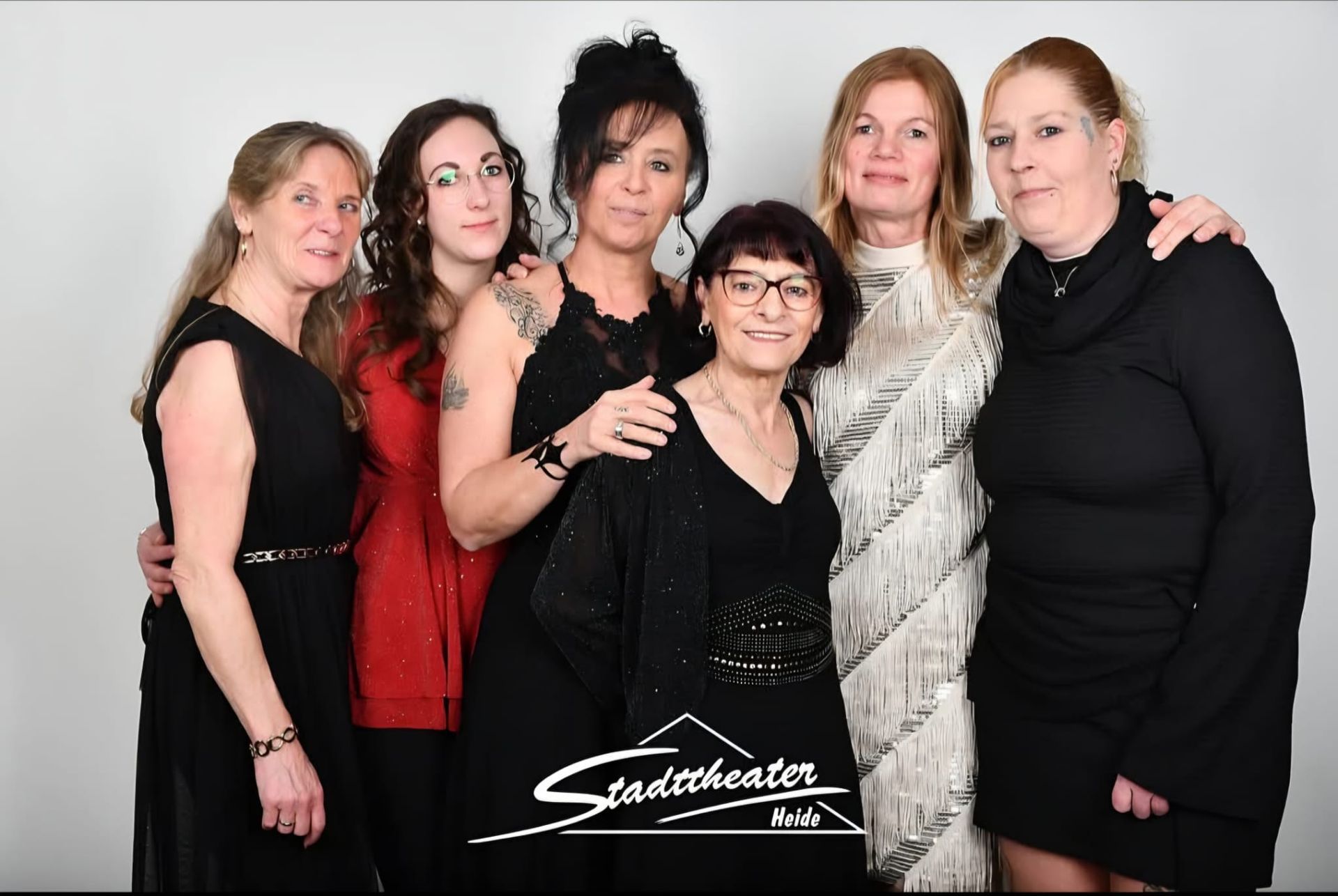 Sechs Frauen posieren gemeinsam vor einem weißen Hintergrund, tragen schwarze und rote Outfits und lächeln.