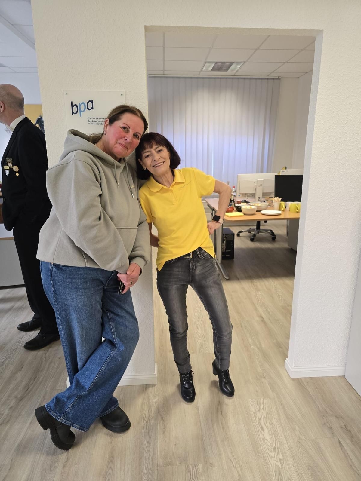 Zwei Arbeitskollegen lehnen lächelnd in einem Büroflur aneinander, einer trägt einen grauen Kapuzenpullover, der andere ein gelbes Hemd.
