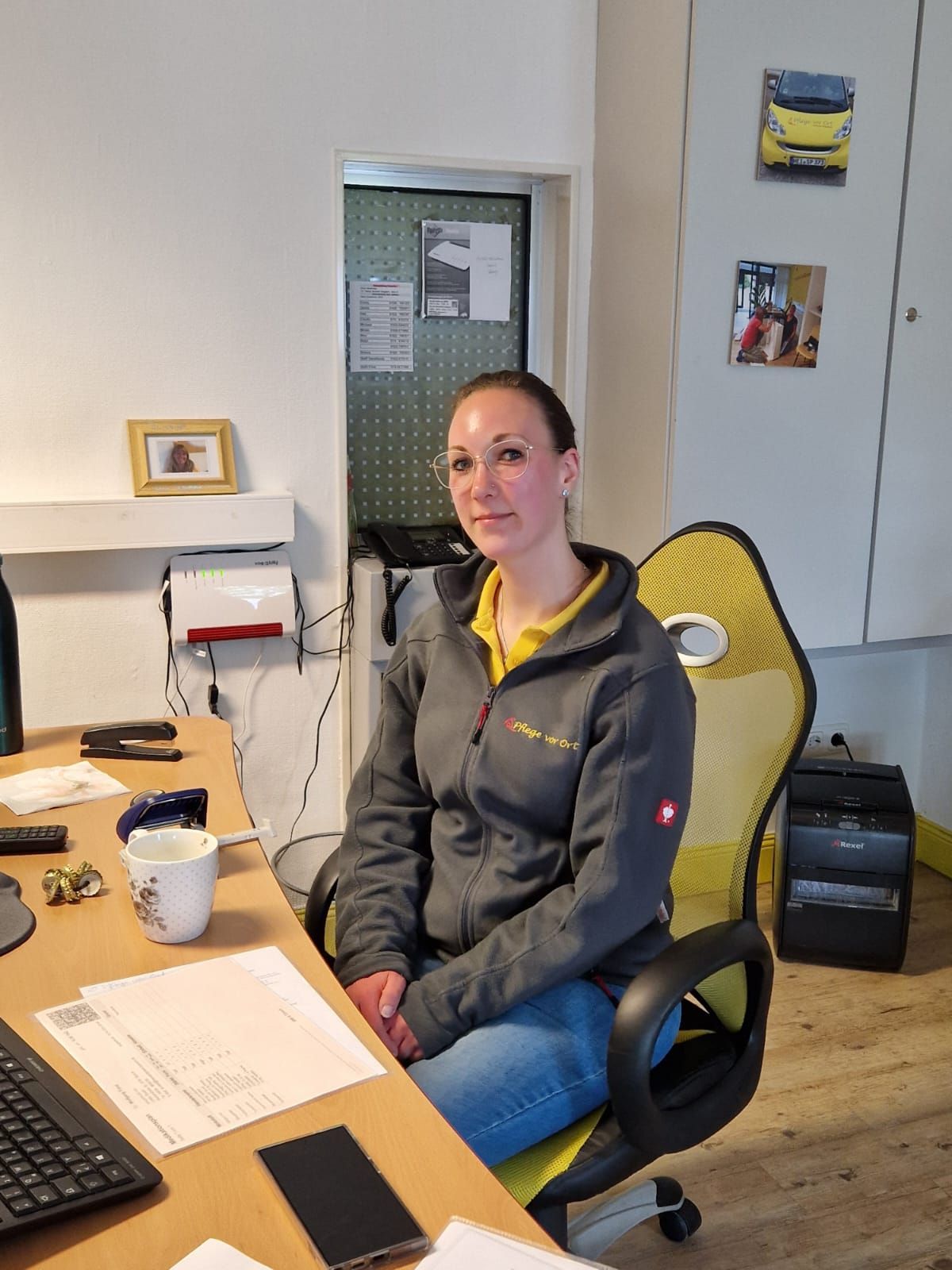 Eine Frau sitzt an einem Schreibtisch in einem Büro; neben ihr liegen Papiere, ein Laptop und eine Kaffeetasse.