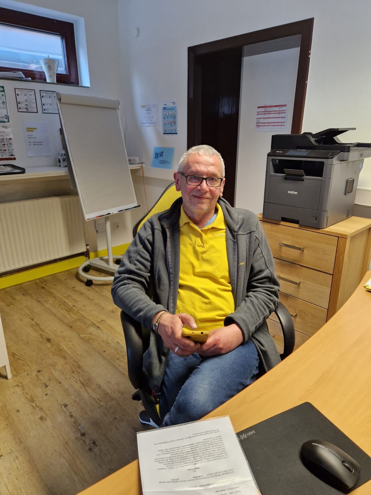 Ein Mann sitzt an einem Schreibtisch in einem Büro. Er trägt ein gelbes Hemd und eine graue Jacke. Hinter ihm befinden sich ein Drucker und ein Whiteboard.