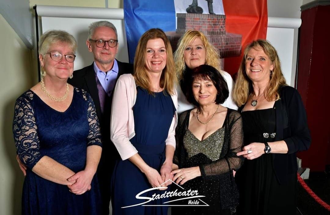 Sechs Personen posieren gemeinsam in einem Innenraum bei einer formellen Veranstaltung, gekleidet in schwarz-blaue Outfits.