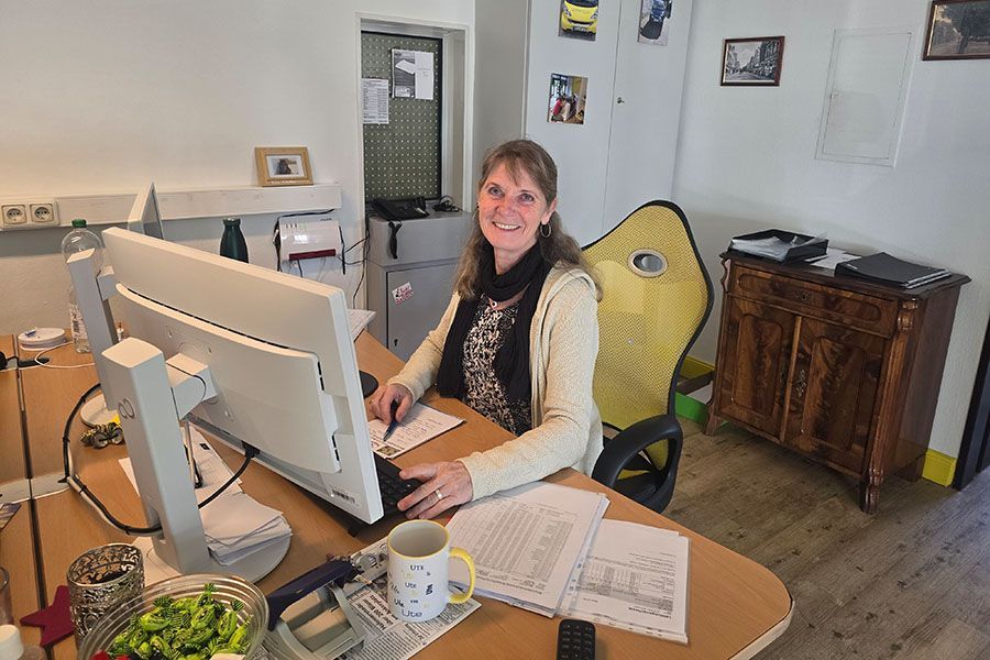 Frau lächelt an einem Schreibtisch mit Computer in ihrem Heimbüro, umgeben von Papieren und einer Kaffeetasse.