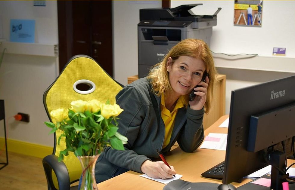 Eine Frau telefoniert am Schreibtisch neben einem Computer und einer Vase mit gelben Blumen.