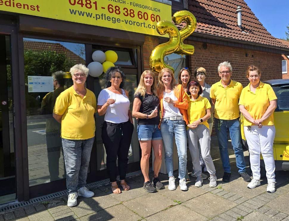 Eine Gruppe von Menschen in gelben Hemden posiert vor einem Schaufenster mit einem Ballonbogen und einer sonnenbeschienenen Straße.