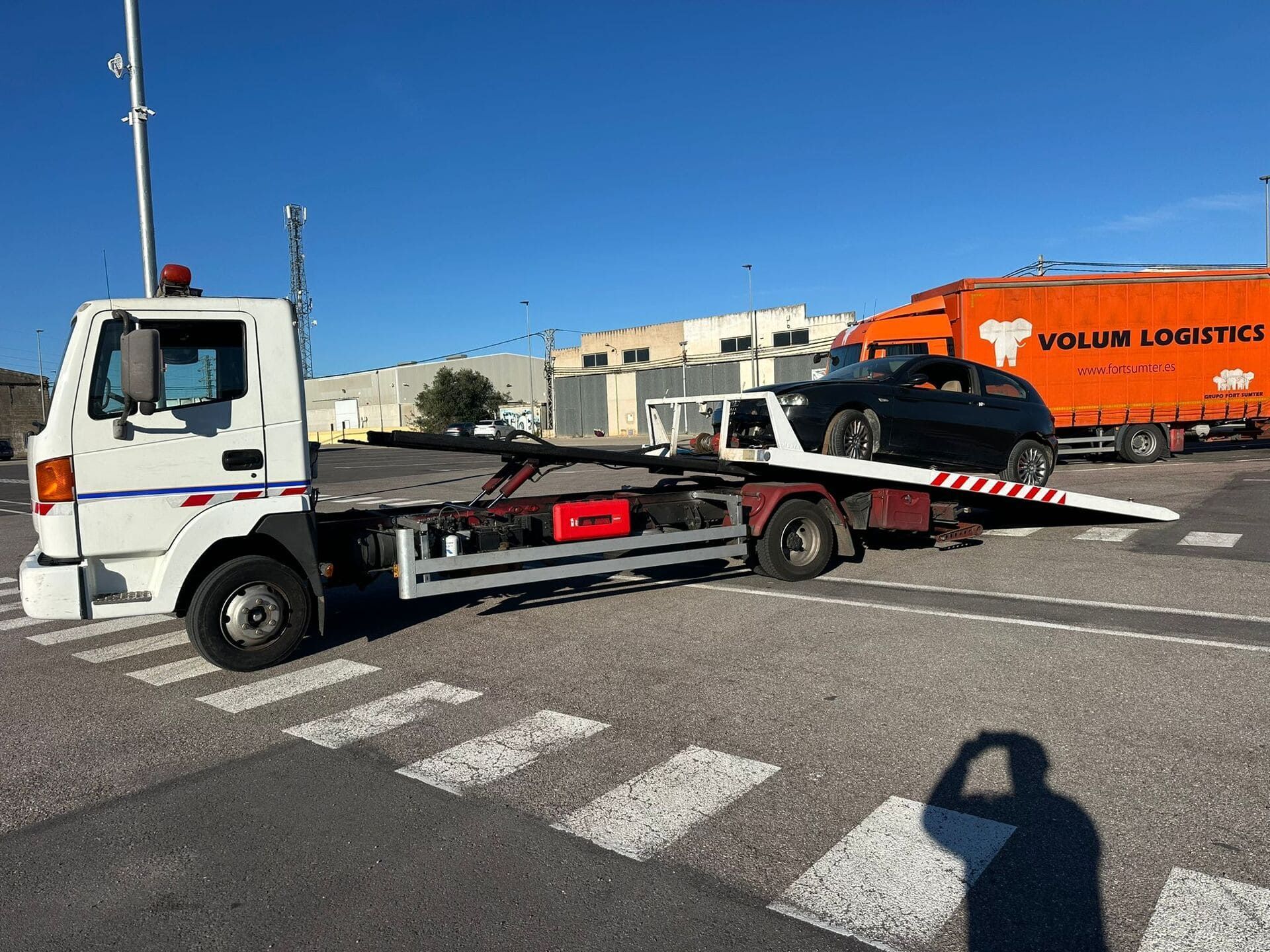 Una grúa blanca en un estacionamiento transportando un automóvil negro, con un gran camión de logística naranja estacionado al fondo.
