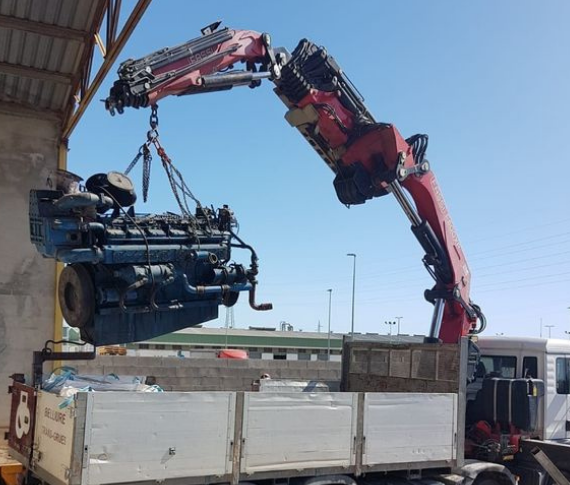 Una grúa montada sobre un camión levanta un gran motor azul de alta resistencia, suspendiéndolo sobre la plataforma del camión bajo un cielo azul despejado.