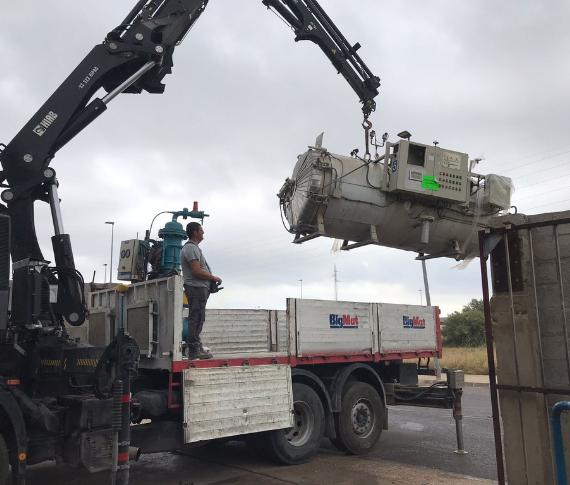 Una grúa levanta un gran transformador eléctrico industrial mientras un trabajador la maneja desde la parte trasera de un camión de plataforma.