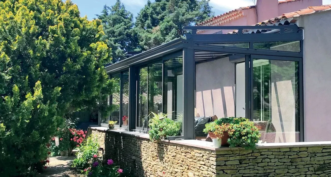 Patio ensoleillé avec pergola en verre, plantes en pot et muret en pierre jouxtant un jardin et une maison.