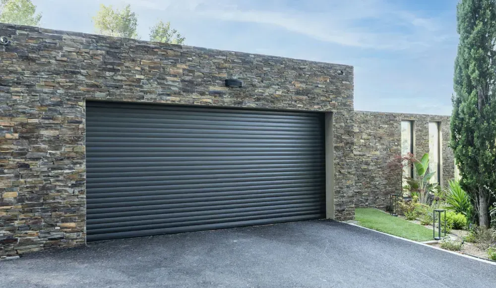 Façade de maison moderne avec une porte de garage sectionnelle sombre, un mur en pierre et une allée sous un ciel bleu
