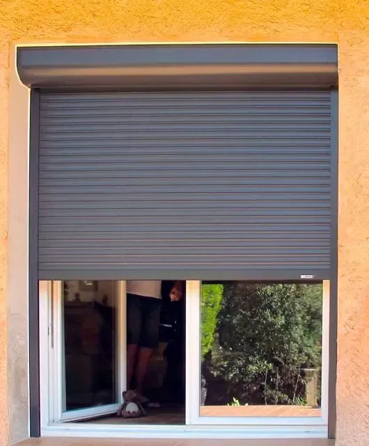 Volet roulant gris partiellement ouvert au-dessus d'une fenêtre coulissante dans un mur extérieur orange