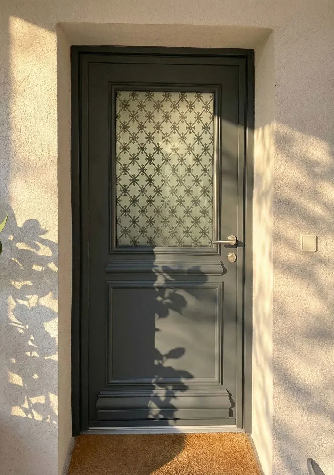 Porte d'entrée grise avec verre dépoli à motifs, encadrée par un enduit beige et des ombres mouchetées.