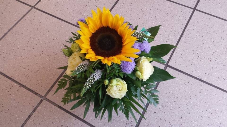 Eine Sonnenblume ist von anderen Blumen auf einem gefliesten Boden umgeben. Foto von der Schöni Blumen und Gartenbau GmbH