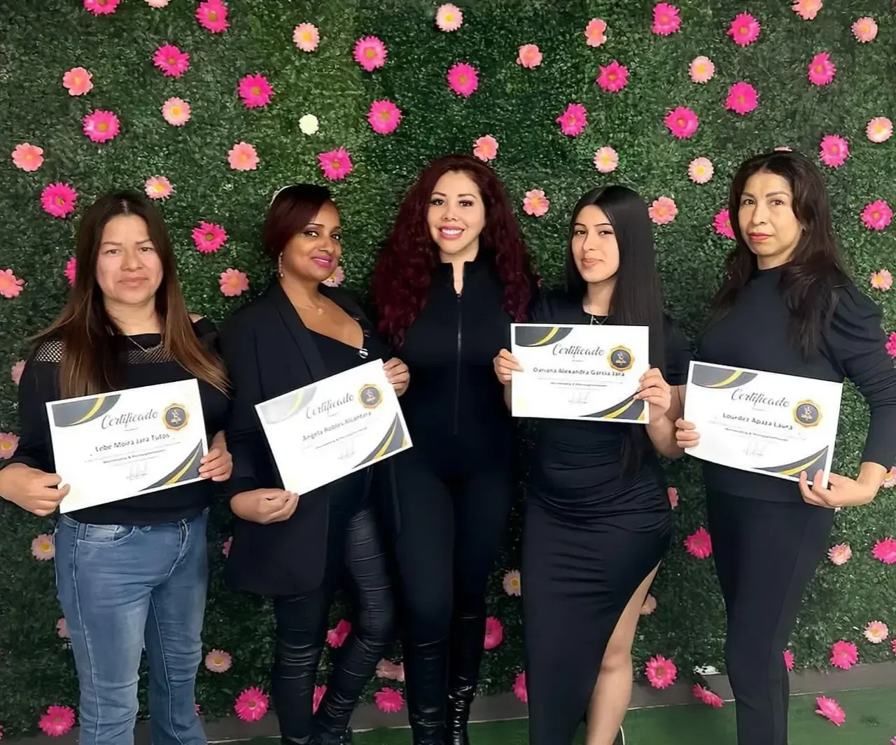 Cinco personas sonríen mientras sostienen certificados frente a una pared decorada con flores rosas y vegetación.