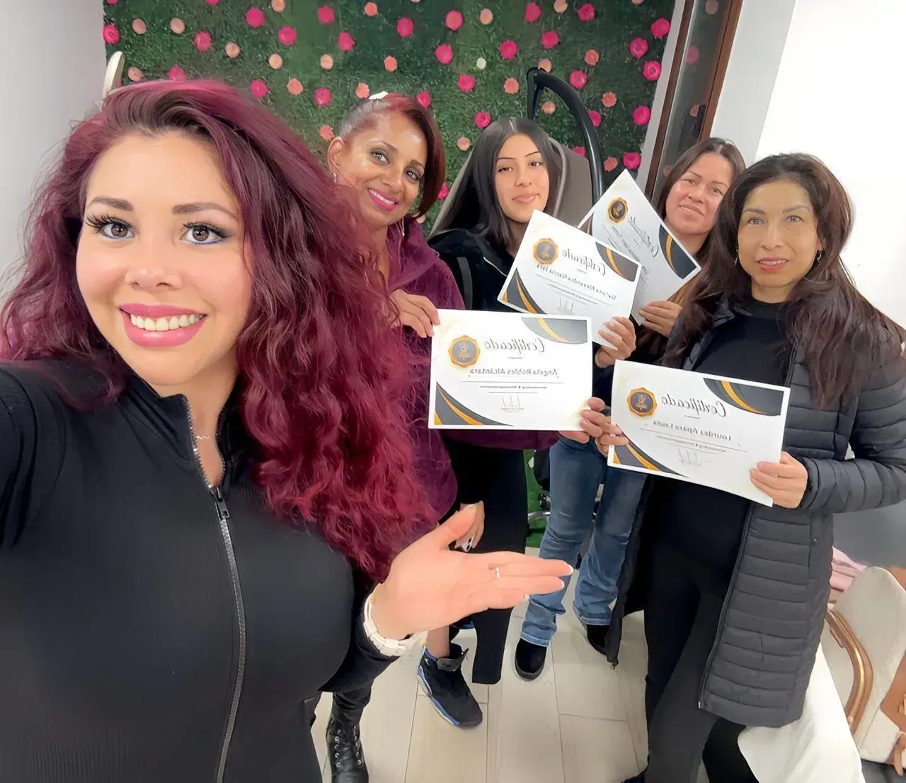 Cinco personas sonriendo y posando con sus certificados en una habitación con una pared decorada con flores.