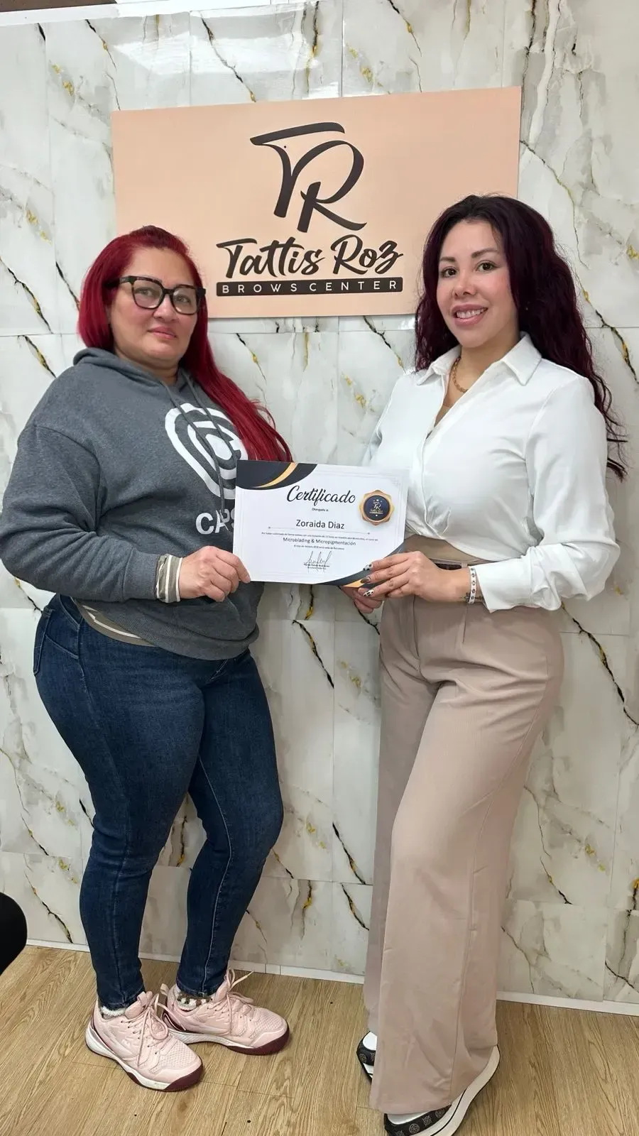 Dos mujeres de pie junto a una pared con un logotipo, sonriendo mientras sostienen un certificado blanco entre ellas.