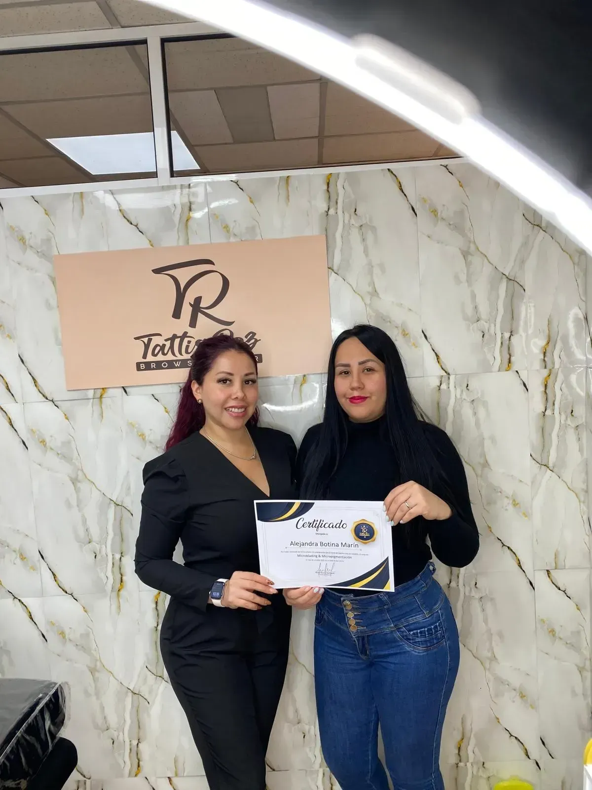 Dos personas sostienen un certificado en un estudio con paredes que imitan el mármol y un letrero comercial.