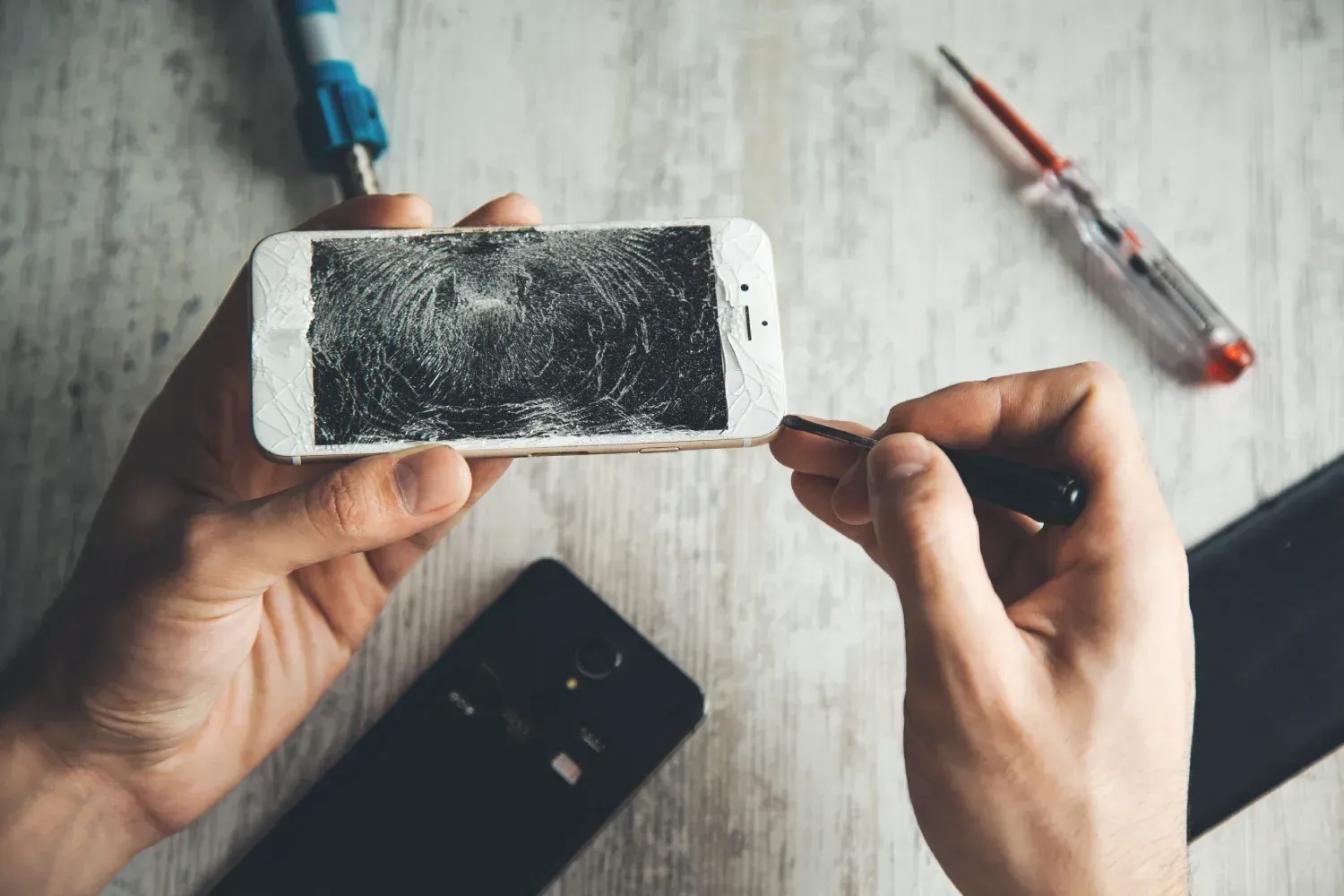 Persona reparando una pantalla de teléfono inteligente dañada con herramientas sobre una superficie de madera.