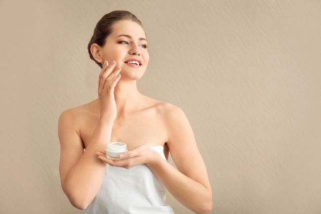 Una mujer envuelta en una toalla se aplica crema en la cara.