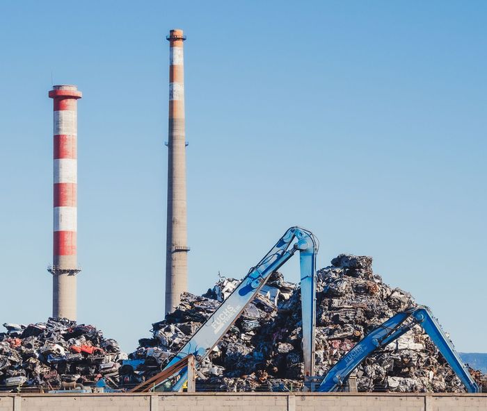 Vertedero industrial con montones de basura, excavadoras azules y dos altas chimeneas bajo un cielo despejado.