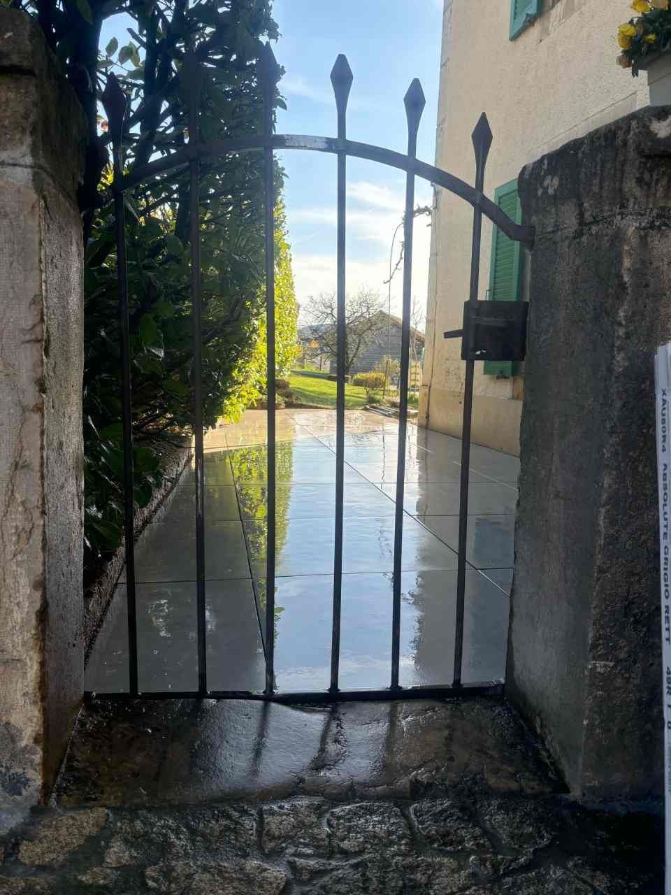 Un portail en fer forgé s'ouvre sur une allée en pierre menant à une maison.