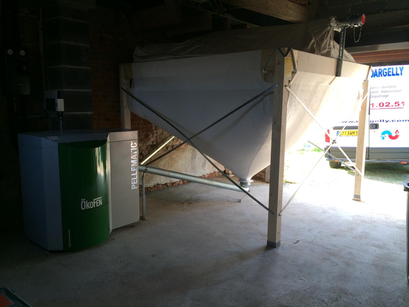 Chaudière à granulés et silo dans une pièce sombre. Chaudière verte et grise, silo blanc sur châssis en bois.