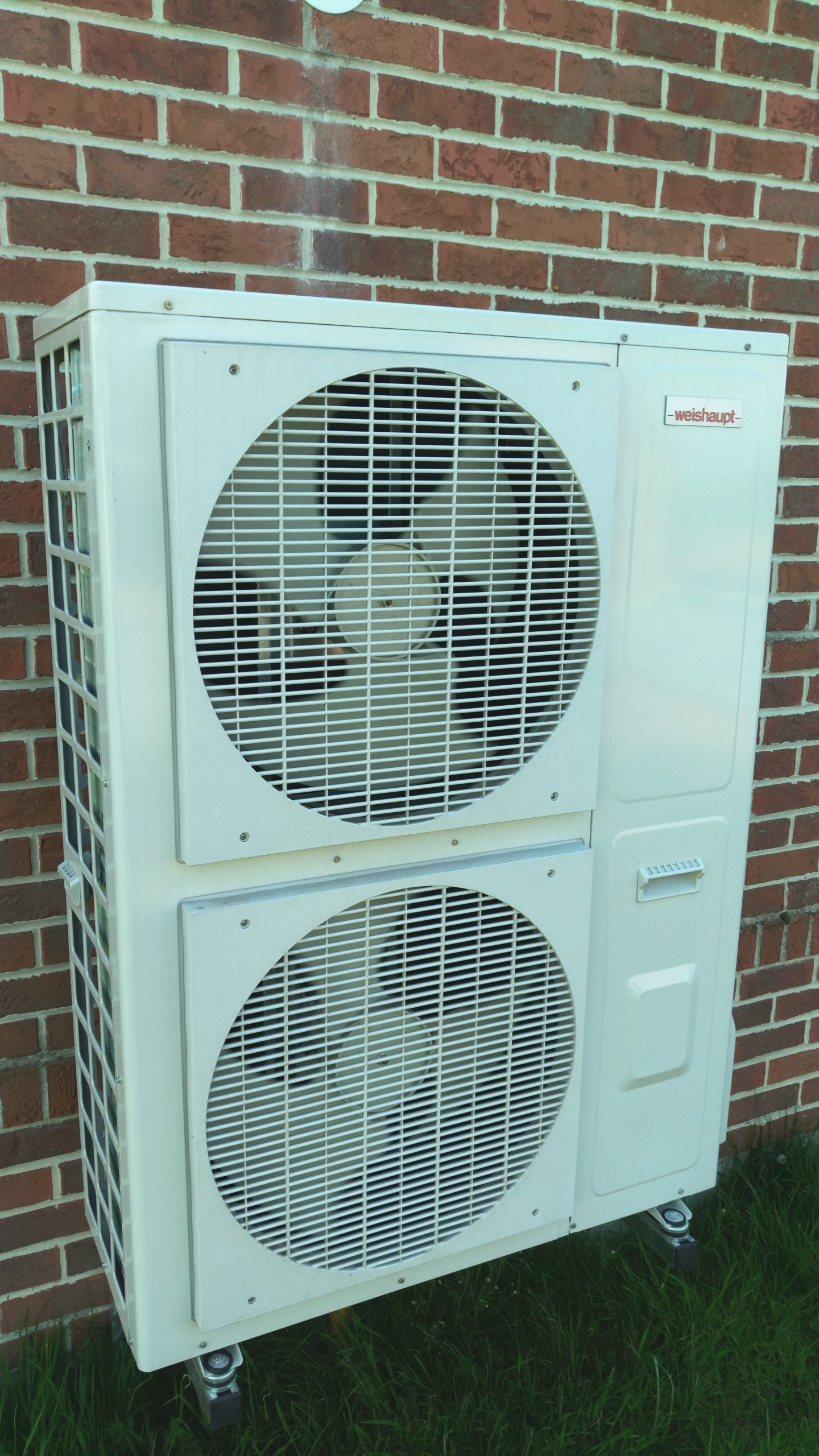 Unité extérieure de pompe à chaleur avec deux ventilateurs, installée contre un mur en briques.