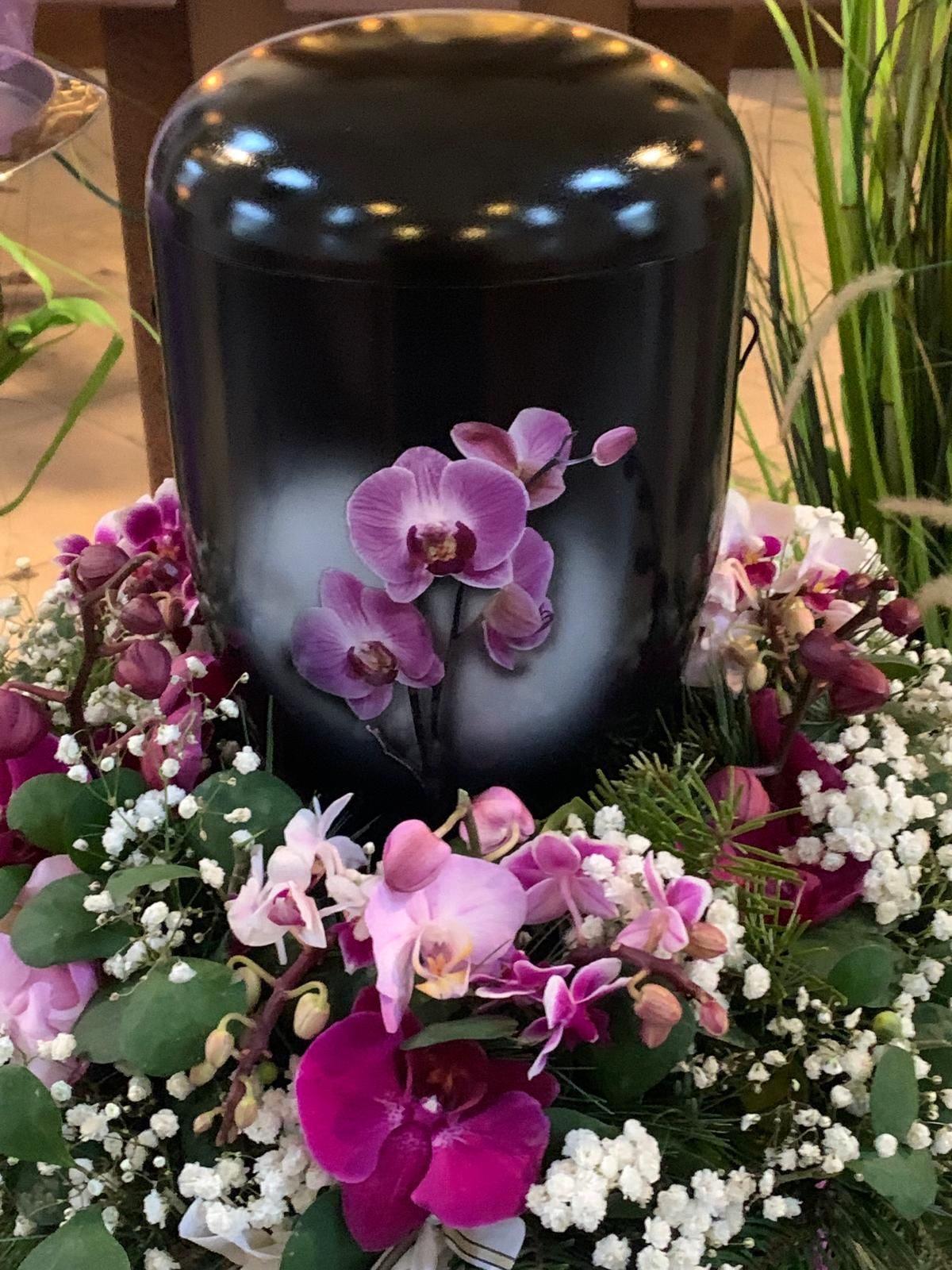 Schwarze Urne mit Orchideenkunst, umgeben von einem Blumenkranz aus Orchideen und Grünpflanzen.