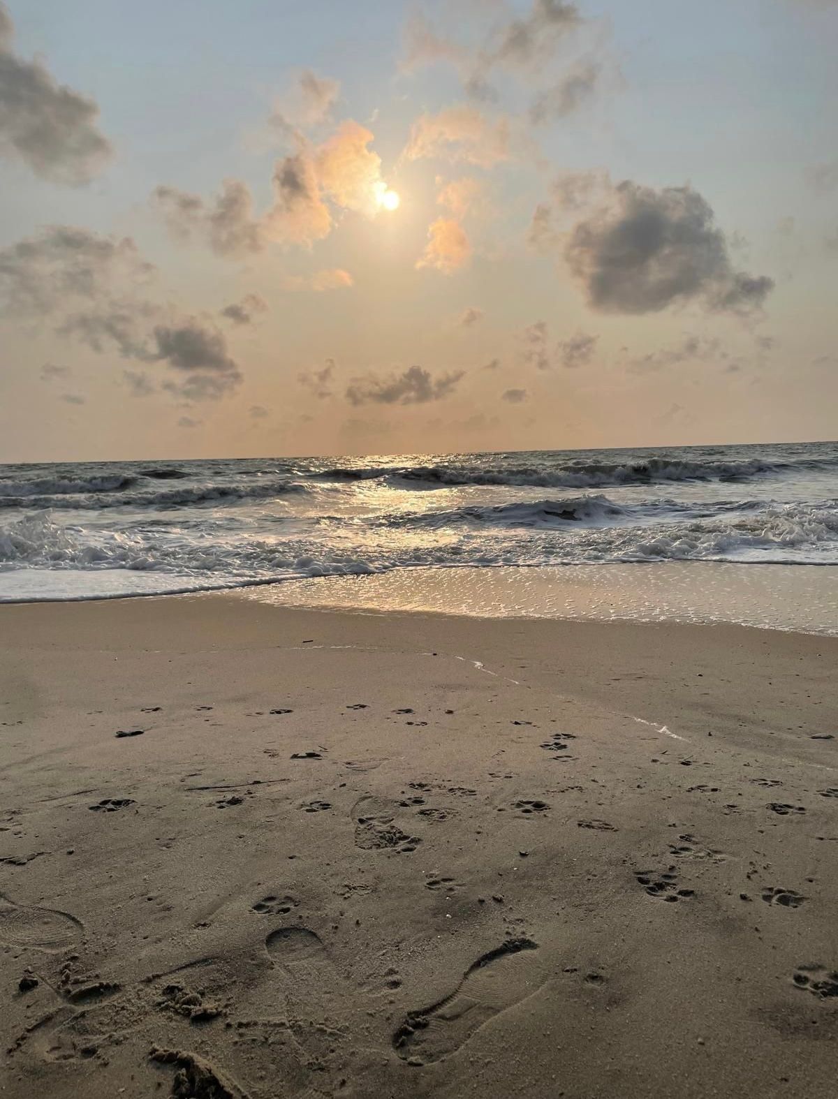 Strand bei Sonnenuntergang, Sonne spiegelt sich im Wasser. Fußspuren im Sand, bewölkter Himmel.