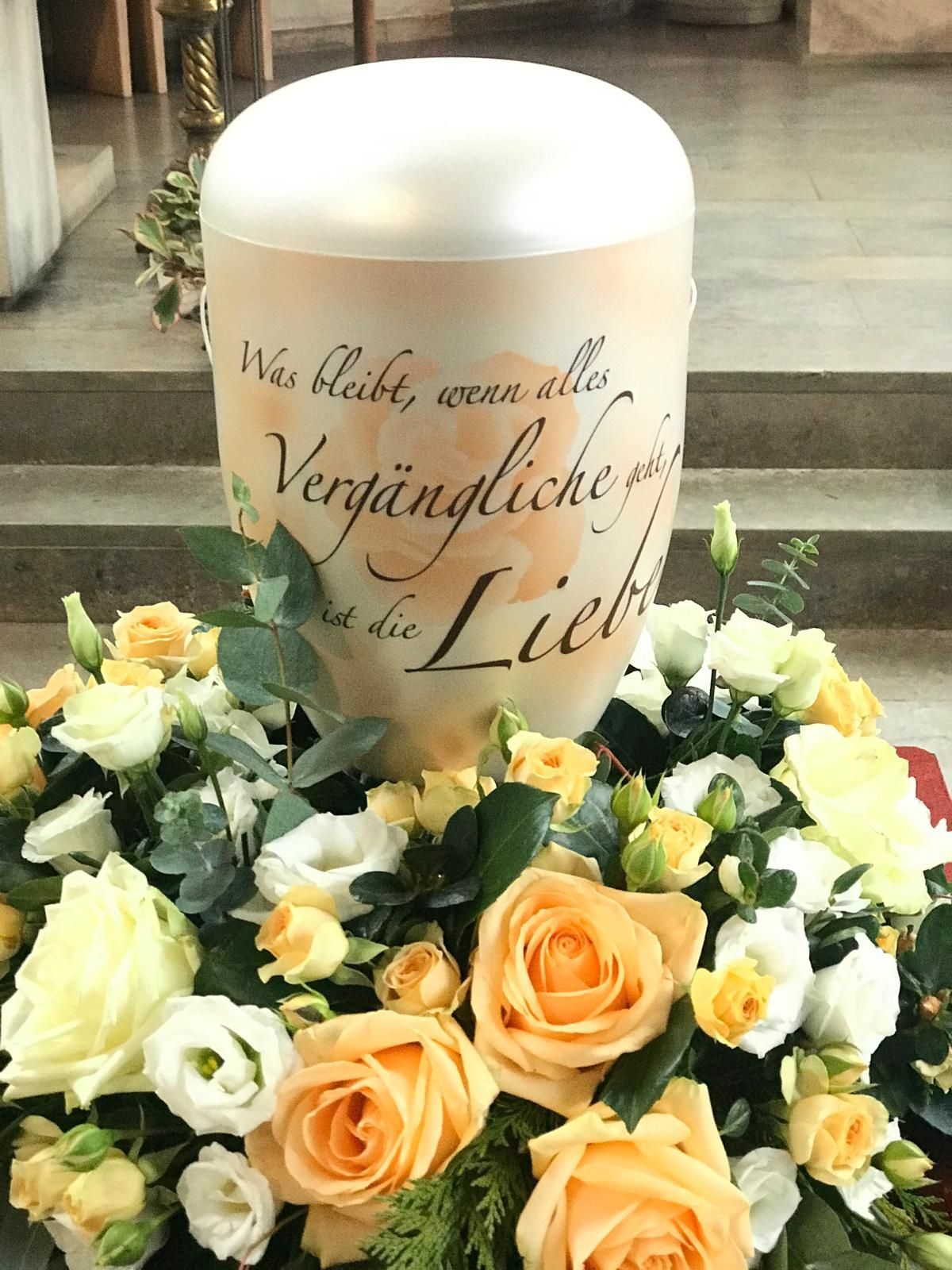 Urne mit Blumenarrangement; Zitat „Liebe“ auf der Urne, mit Rosen und anderen Blumen, im Innenbereich.