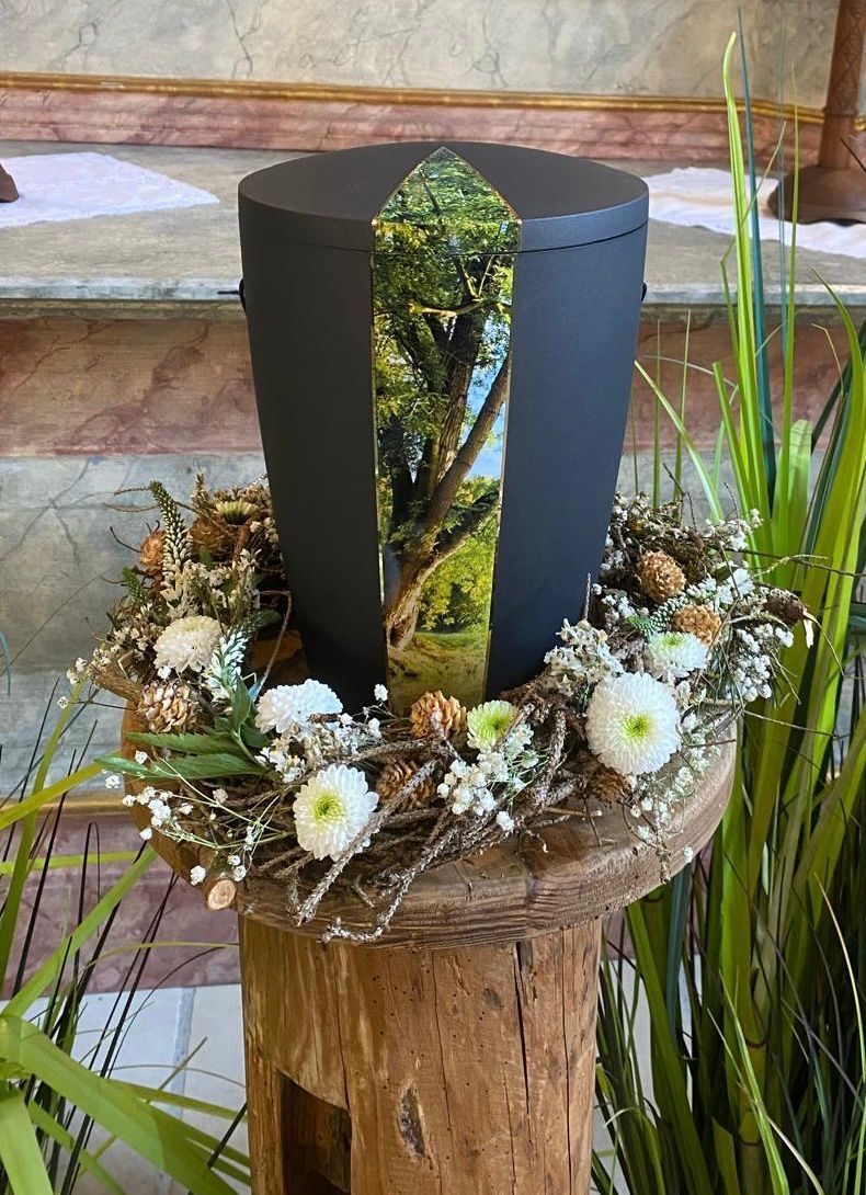 Schwarze Urne mit malerischem Bild, umgeben von einem Blumenkranz, auf einem Holzsockel.
