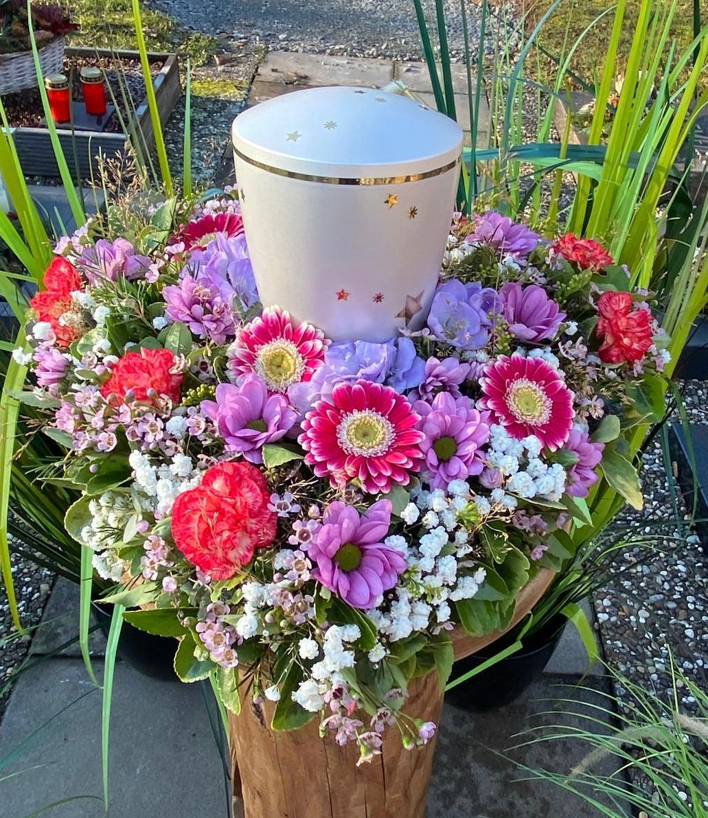 Urne umgeben von bunten Blumen: rot, lila, rosa. Draußen, in einer Gartenumgebung.