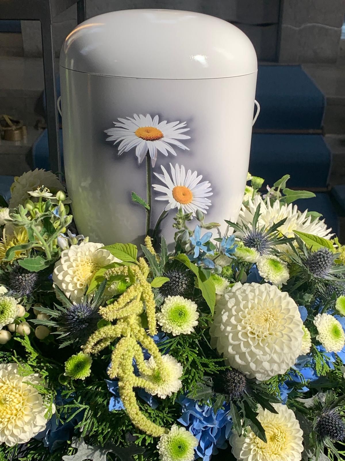 Weiße Urne mit Gänseblümchenmuster, umgeben von einem Blumenarrangement aus weißen, blauen und grünen Blumen.