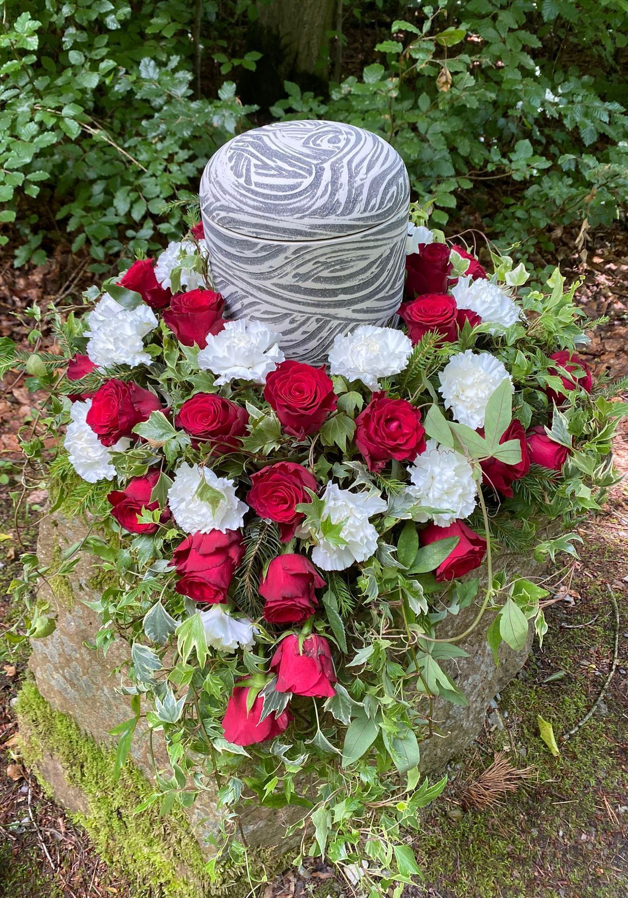 Eine Urne, umgeben von einem Kranz aus roten Rosen und weißen Nelken auf einem moosigen Felsen im Freien.
