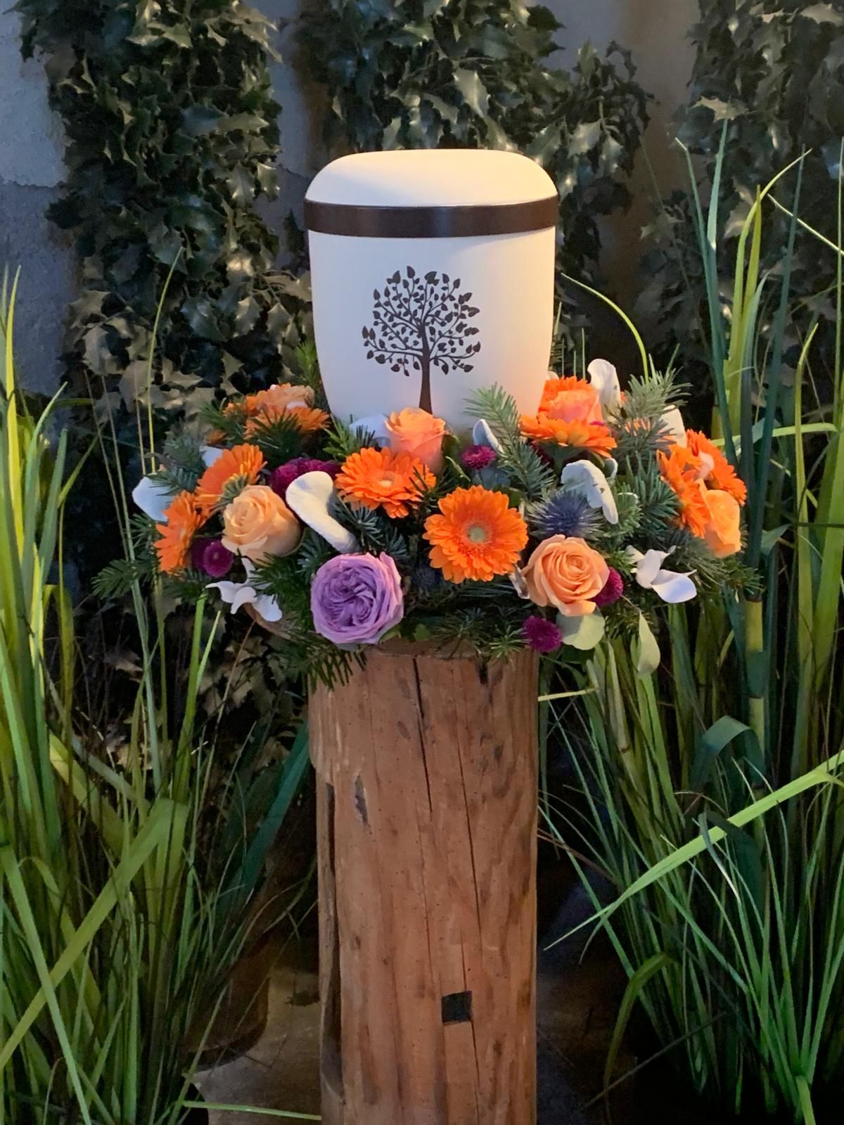 Urne mit Baummotiv auf einem Blumenarrangement auf einem Holzpfosten vor grünem Hintergrund.