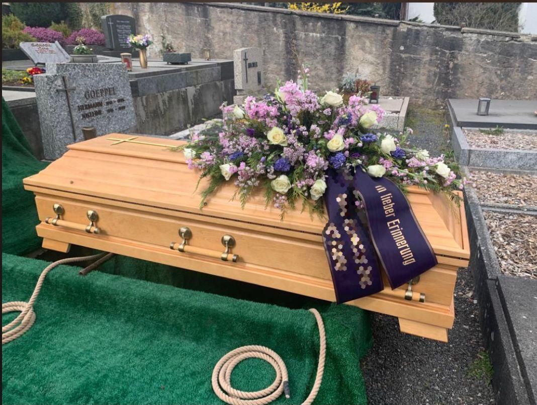 Holzsarg mit Blumenarrangement und Band auf einem Friedhof.