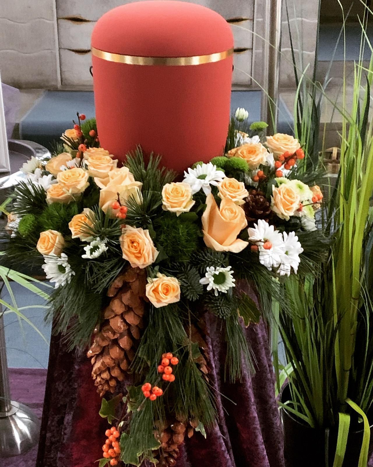 Rote Urne mit Blumenarrangement aus Rosen, Gänseblümchen, Beeren und Grünpflanzen.