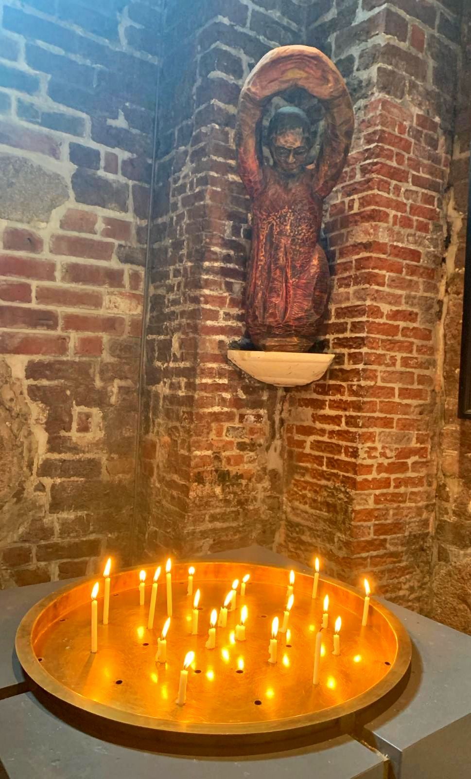 Eine Holzstatue hält eine Schale über einem Kerzentablett, das in einem warm beleuchteten Raum vor einer Ziegelwand steht.