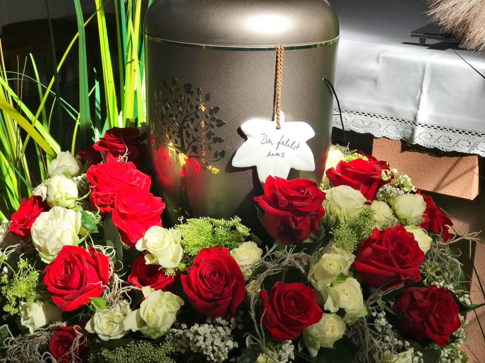 Eine Urne, umgeben von roten Rosen und weißen Blumen, mit einem blattförmigen Anhänger, wahrscheinlich bei einer Gedenkstätte.
