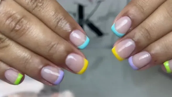 Manos con manicura francesa en tonos pastel de menta, amarillo y lila, juntas sobre un fondo gris.