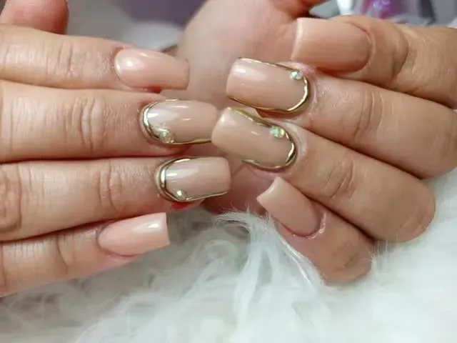 Manos con uñas color nude y puntas doradas, unidas sobre una tela blanca y esponjosa.