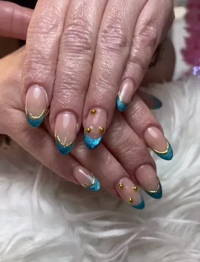 Primer plano de unas manos con uñas color nude y puntas francesas turquesa y doradas, adornadas con pequeñas gemas doradas.