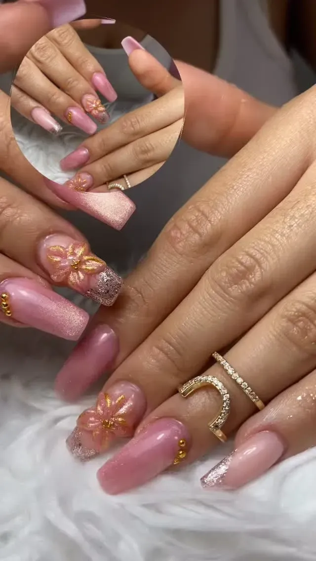 Manos con uñas largas de color rosa bien cuidadas y anillos de oro, con uñas decoradas con detalles florales brillantes.