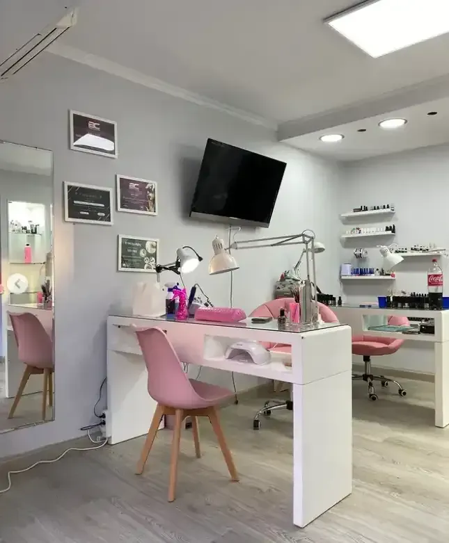 Interior de salón de uñas con detalles en rosa, mesas de manicura, sillas, lámparas y televisor montado en la pared.