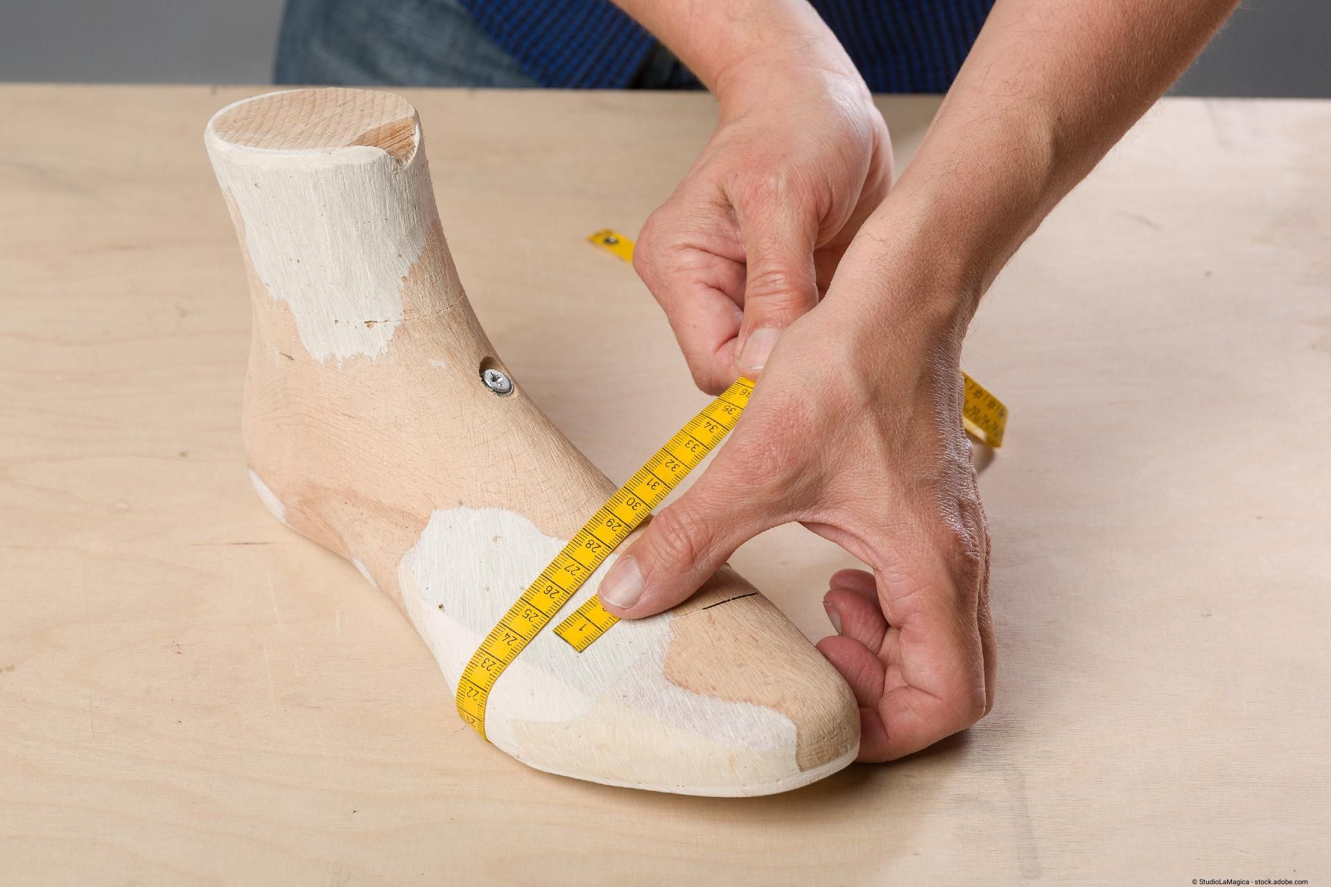 Hände messen mit einem Maßband den Umfang eines Fußes mit angelegtem Gipsabdruck auf einem Tisch.