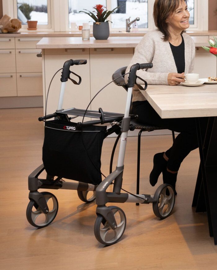 Seniorin sitzt am Tisch mit Rollator in der Küche und trinkt eine Tasse Kaffee.