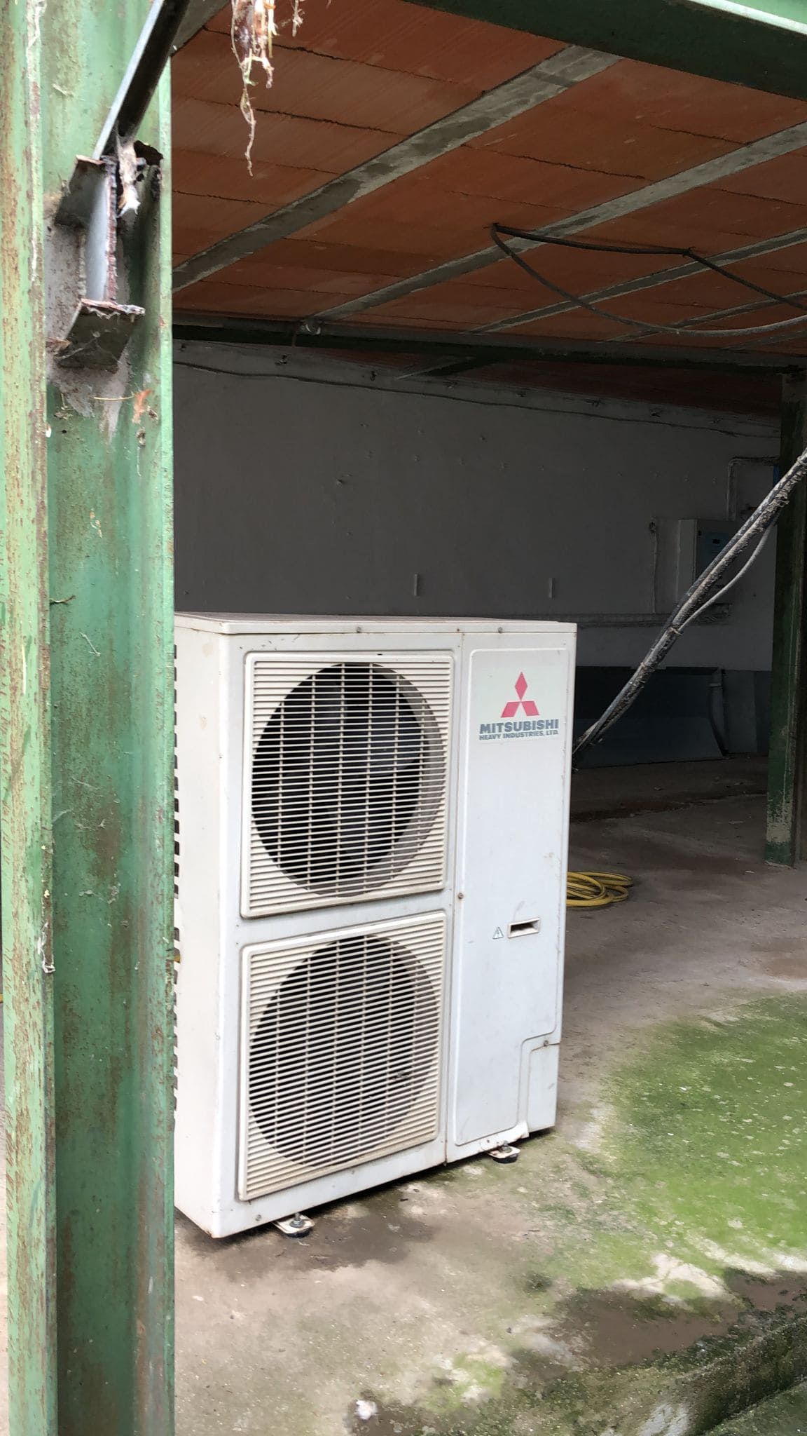 Una unidad exterior de aire acondicionado Mitsubishi de color blanco se encuentra sobre un suelo de hormigón bajo un cobertizo rústico y sombreado.