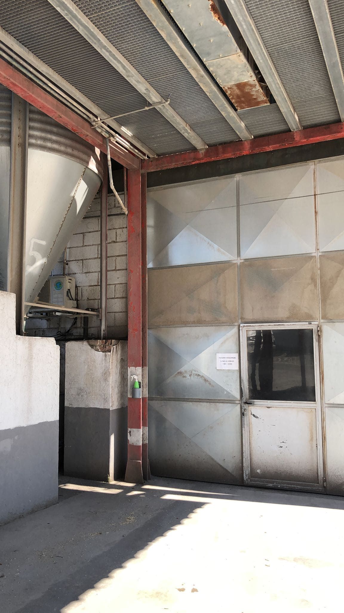 Una estructura industrial con una pared metálica, una puerta y una tolva cónica bajo un techo de chapa ondulada, a plena luz del día.